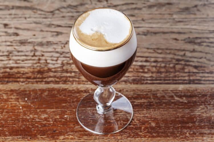 A lgith brown coffee with generous foam in a fluted clear glass on a brown table in Austria.